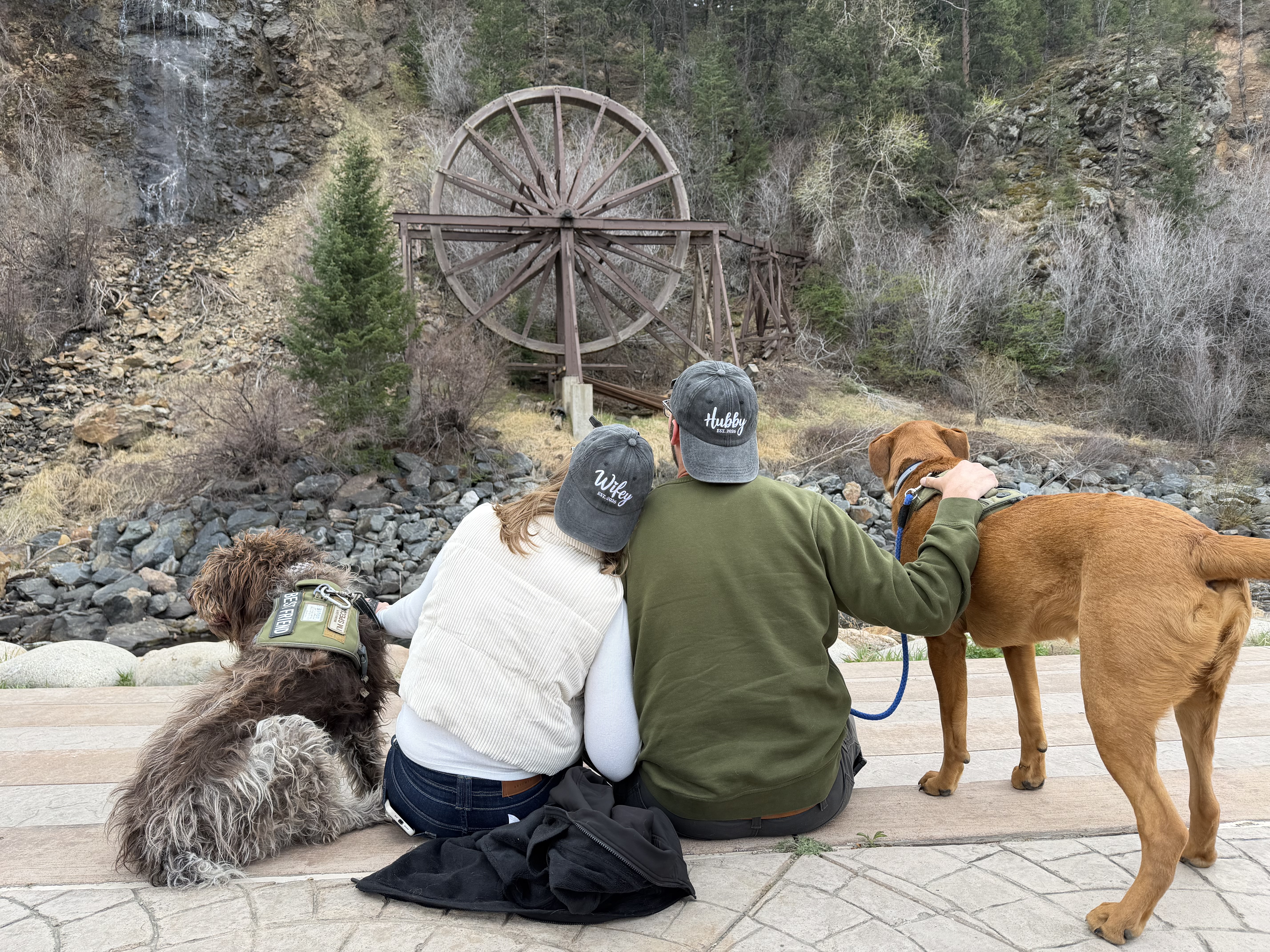 Jamie Watz and Evan Watz seated with their two dogs near a waterfall and waterwheel